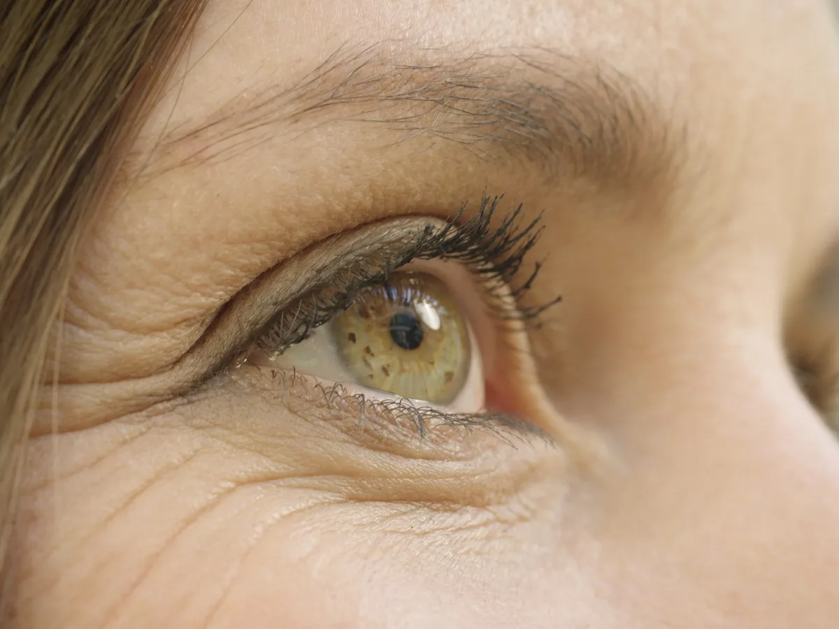 Nahaufnahme eines Auges: grünlich-braune Iris, Lidrand und Wimpern im Detail, dahinter weich verschwommen die Gesichtshaut